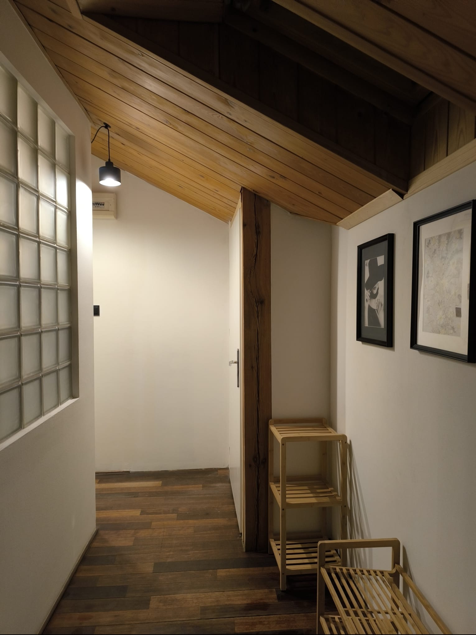 Korytarz z drewnianym sufitem i podłogą. Na ścianie szklane luksfery i grafiki w czarnych ramkach. Widoczny drewniany słupek i małe drewniane regały.