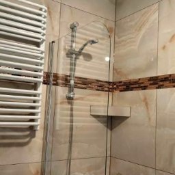 Dom na poziomie - Wykończenia wnętrz Łukasz Burzyński - Wykończona łazienka z beżowymi płytkami imitującymi marmur, kabiną prysznicową ze szklanym panelem i grzejnikiem drabinkowym. Widoczny detal dekoracyjny w postaci mozaiki.