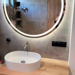 Dom na poziomie - Wykończenia wnętrz Łukasz Burzyński - Nowoczesna łazienka z okrągłym lustrem LED, białą umywalką nablatową na drewnianym blacie i szarą płytką. Widoczne elementy armatury i detale wykończenia.