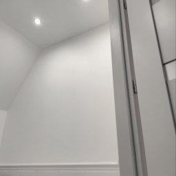 Dom na poziomie - Wykończenia wnętrz Łukasz Burzyński - Białe wnętrze po remoncie: ściana z panelami, sufit z dwoma punktowymi lampami LED, widok przez otwarte, białe drzwi.