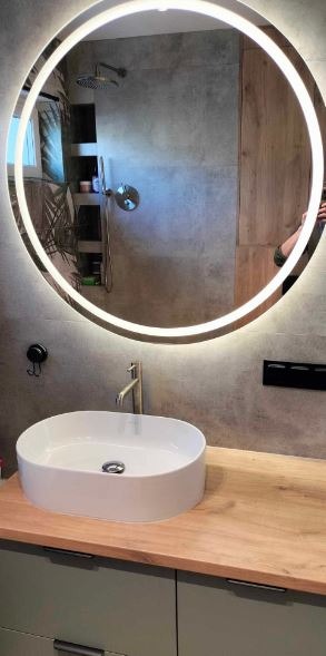 Nowoczesna łazienka z okrągłym lustrem z podświetleniem LED, białą umywalką nablatową i szarą glazurą. Widoczne elementy armatury i drewniane akcenty.