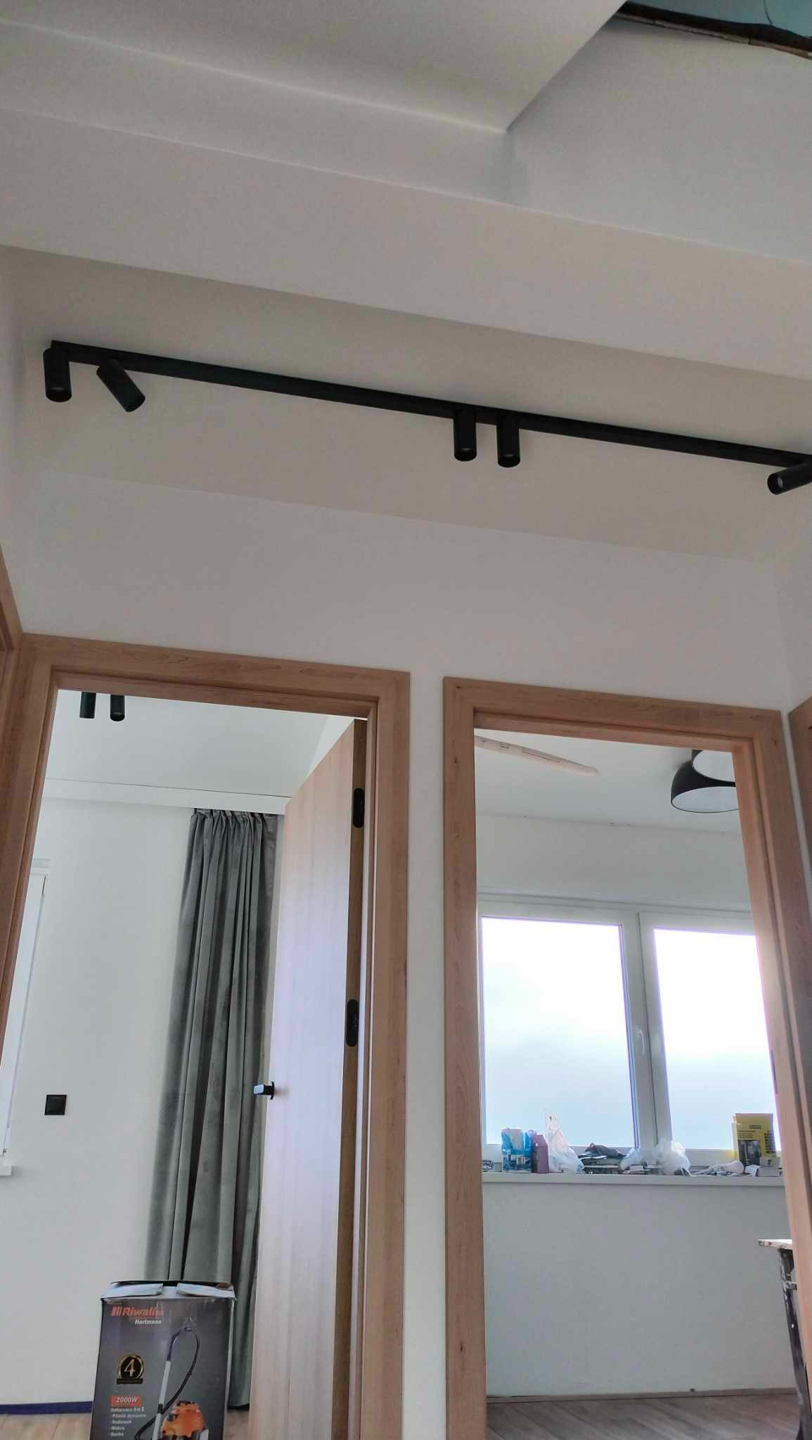 Zamontowane czarne oświetlenie szynowe sufitowe w korytarzu, widoczne dwie pary drewnianych drzwi, wnętrze pokoi i odkurzacz w rogu. Minimalistyczny design.