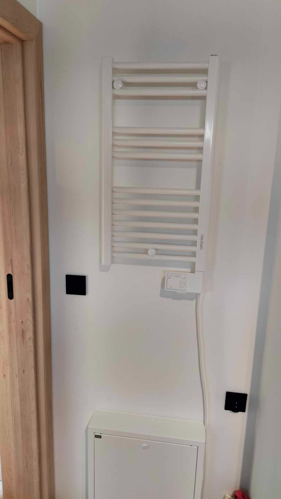 Biały, drabinkowy grzejnik łazienkowy Atlantic zamontowany na białej ścianie, obok drewnianej ościeżnicy. Widoczne czarne włączniki i biała skrzynka pod grzejnikiem.