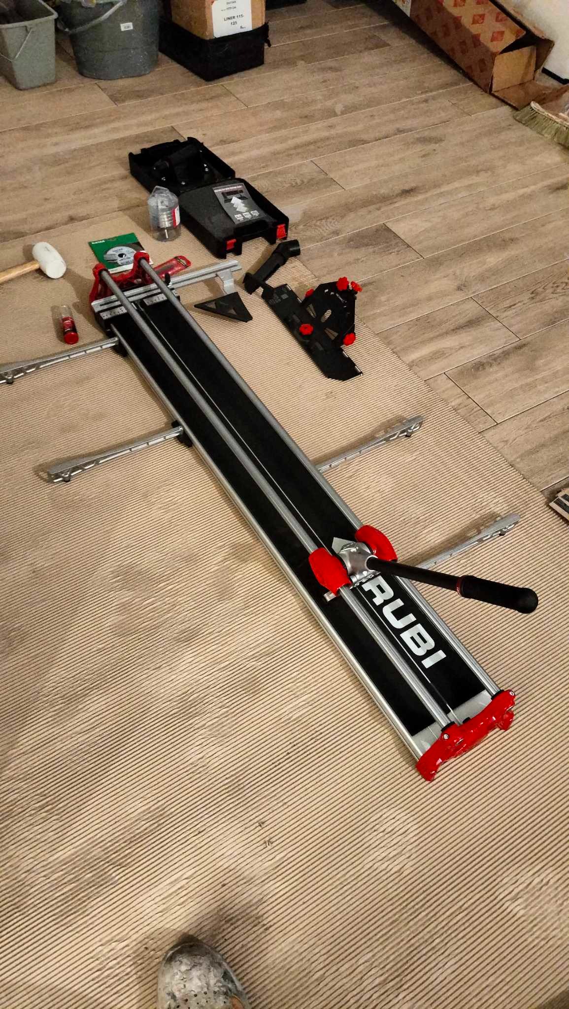 Profesjonalna przecinarka do płytek Rubi, rozłożona na podłodze z widocznymi akcesoriami: młotek gumowy, opakowanie z tarczą, pudełko i pojemnik z wodą.