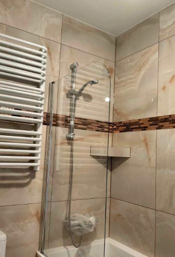 Wykończona łazienka z beżowymi płytkami imitującymi marmur, kabiną prysznicową ze szklanym panelem i grzejnikiem drabinkowym. Widoczny detal dekoracyjny w postaci mozaiki.