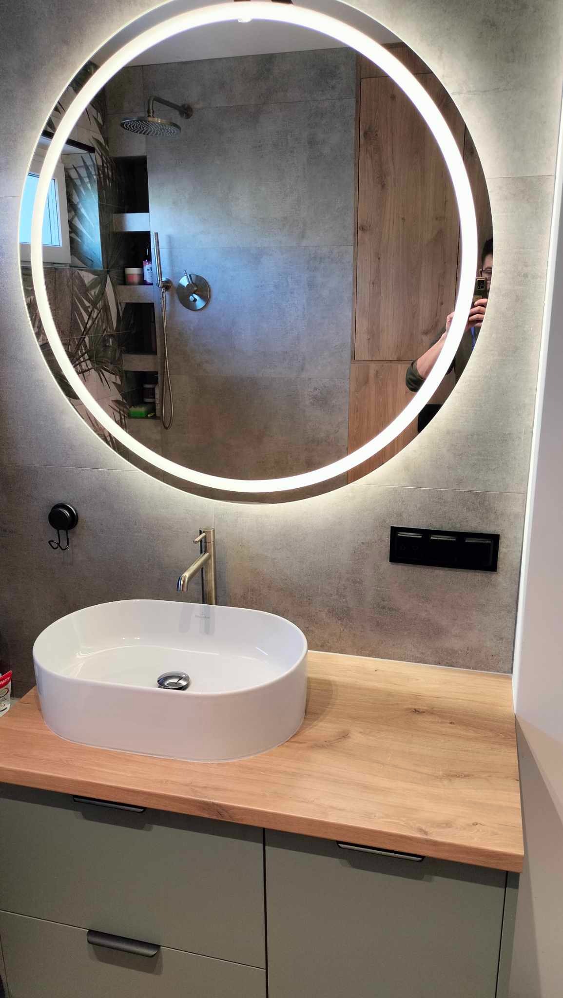 Nowoczesna łazienka z okrągłym lustrem LED, białą umywalką nablatową na drewnianym blacie i szarą płytką. Widoczne elementy armatury i detale wykończenia.