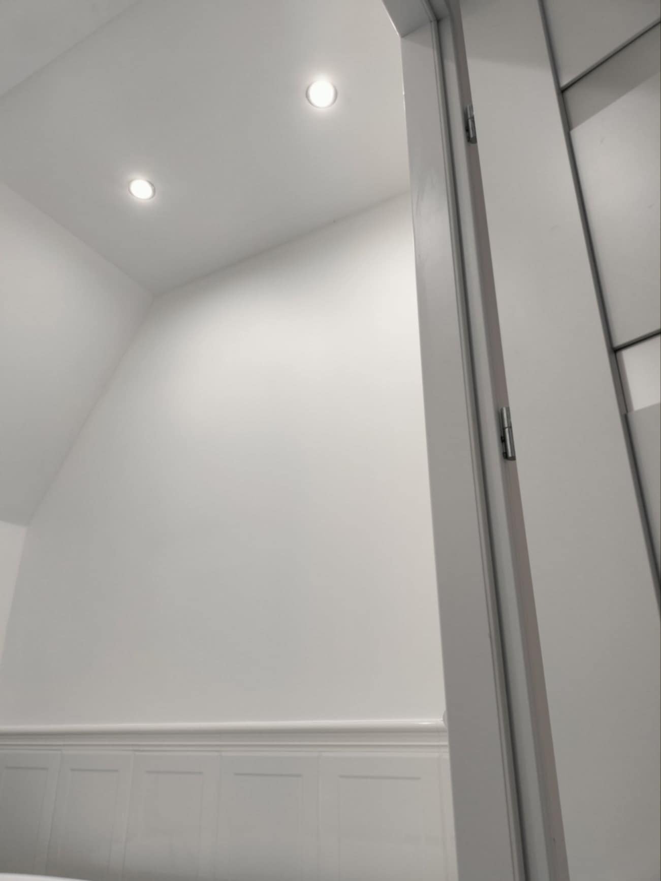 Białe wnętrze po remoncie: ściana z panelami, sufit z dwoma punktowymi lampami LED, widok przez otwarte, białe drzwi.