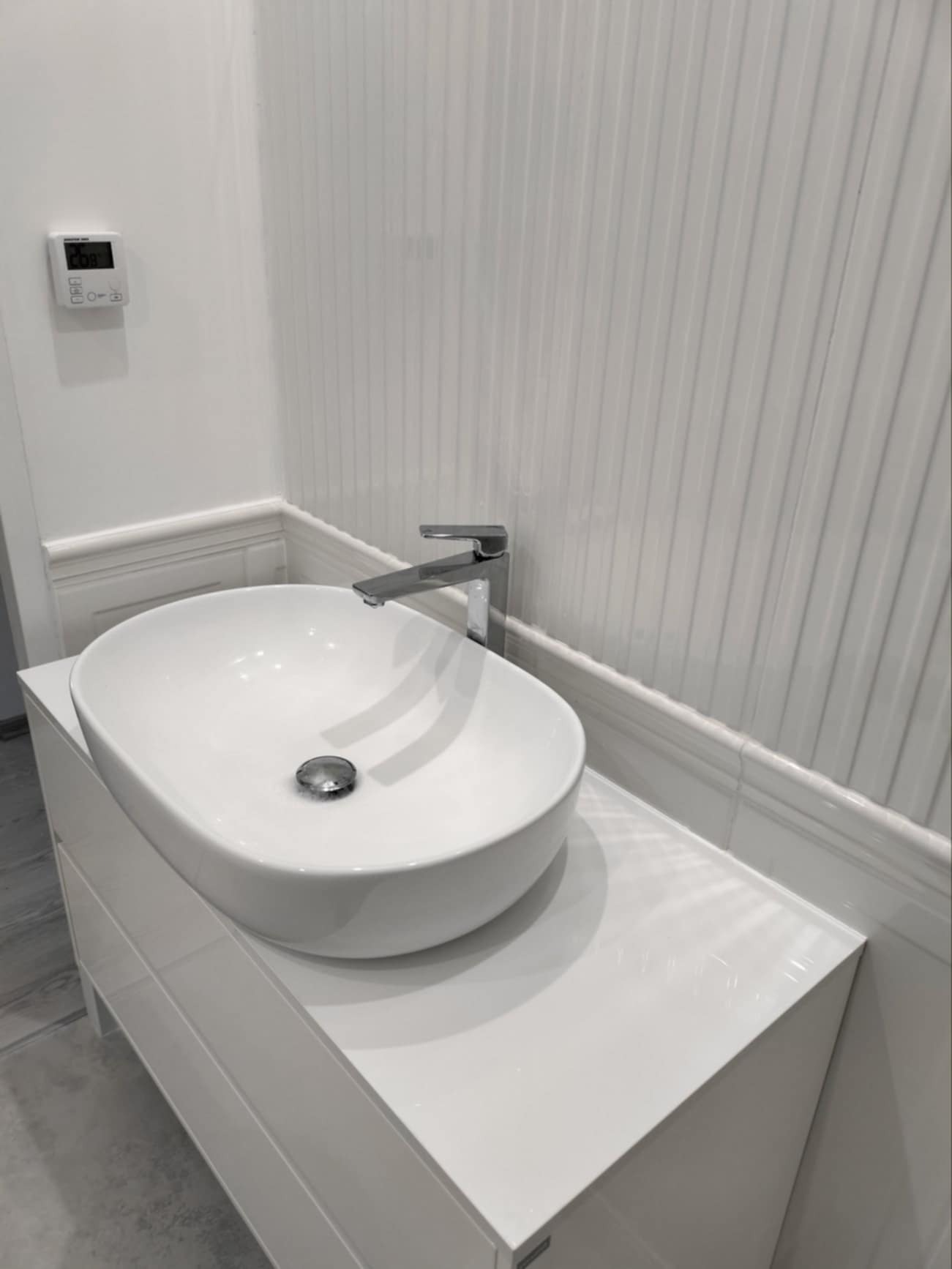 Biała, owalna umywalka nablatowa z chromowaną baterią na szafce w nowoczesnej łazience z panelami ściennymi. Minimalistyczny design, czyste linie.