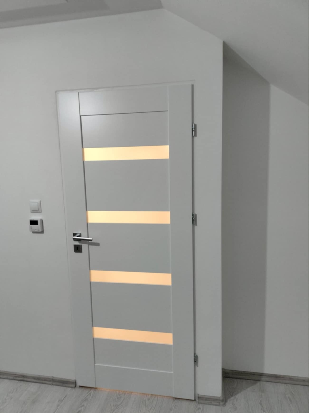 Białe drzwi wewnętrzne z poziomymi przeszkleniami, zamontowane w jasnym pomieszczeniu ze skośnym sufitem, widoczne gniazdka elektryczne na ścianie.