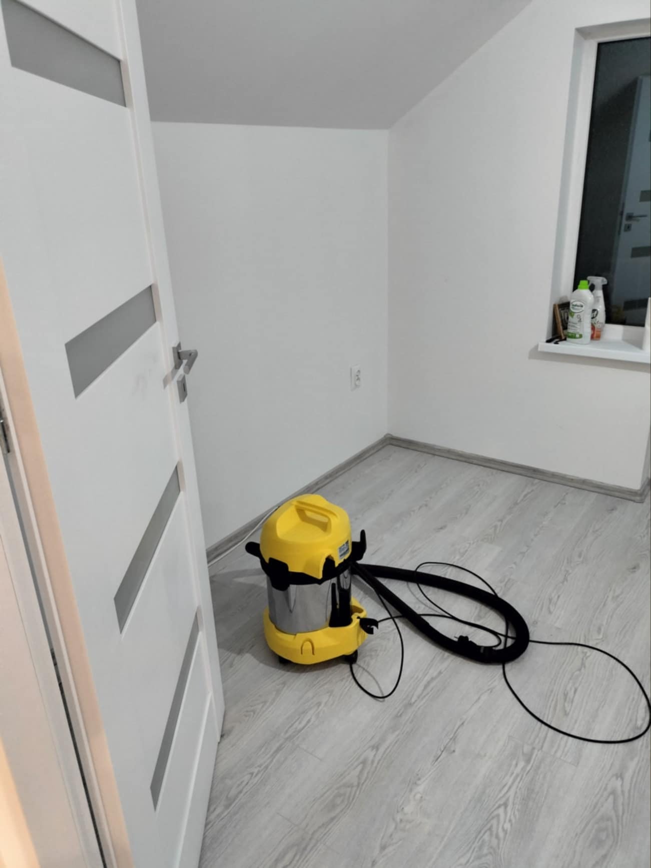 Wnętrze pokoju po remoncie z nową podłogą i odkurzaczem przemysłowym. Widoczna biała ściana, okno i drzwi z szarymi wstawkami. Oświetlenie dzienne.