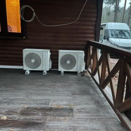 INSTAL-BUD KONRAD ZALECH - Dwa klimatyzatory na drewnianym tarasie domu z brązową elewacją. Na zewnątrz widoczny biały samochód dostawczy i drzewa.