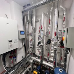 INSTAL-BUD KONRAD ZALECH - Węzeł cieplny z pompą ciepła Panasonic Aquarea Smart Cloud, licznymi rurami z izolacją, manometrami i zaworami w pomieszczeniu technicznym.