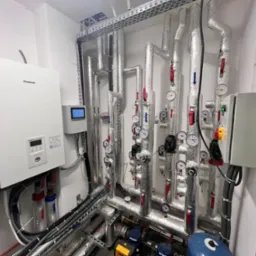 Węzeł cieplny z pompą ciepła Panasonic Aquarea Smart Cloud, licznymi rurami z izolacją, manometrami i zaworami w pomieszczeniu technicznym.