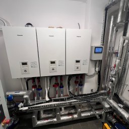 INSTAL-BUD KONRAD ZALECH - Trzy białe pompy ciepła Panasonic zamontowane na ścianie z rurami, z widocznym panelem sterowania i elementami instalacji. Rury zaizolowane, z manometrami.