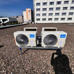 INSTAL-BUD KONRAD ZALECH - Dwa klimatyzatory Midea, zamontowane na metalowej konstrukcji, na dachu pokrytym drobnym żwirem. W tle budynki w słoneczny dzień. Widoczny cień osoby robiącej zdjęcie.