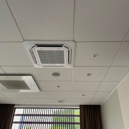 INSTAL-BUD KONRAD ZALECH - Kasetonowa klimatyzacja sufitowa z widoczną kratką wentylacyjną i oświetleniem punktowym. Okno z żaluzjami i widokiem na zieleń.