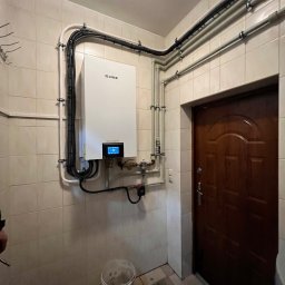 INSTAL-BUD KONRAD ZALECH - Zamontowany biały piec grzewczy marki Gree na kafelkowej ścianie, z widocznymi rurami i przewodami, w pomieszczeniu z brązowymi drzwiami i wiadrem na podłodze.