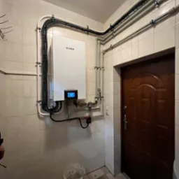 Zamontowany biały piec grzewczy marki Gree na kafelkowej ścianie, z widocznymi rurami i przewodami, w pomieszczeniu z brązowymi drzwiami i wiadrem na podłodze.