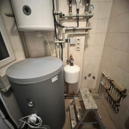 INSTAL-BUD KONRAD ZALECH - Szary zbiornik buforowy z białym zasobnikiem wody i instalacją hydrauliczną na tle beżowych płytek. Obok drabina.