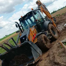 Pomarańczowa koparko-ładowarka CASE w błotnistym terenie, widok z dołu, ślady opon w ziemi, pola w tle pod pochmurnym niebem.