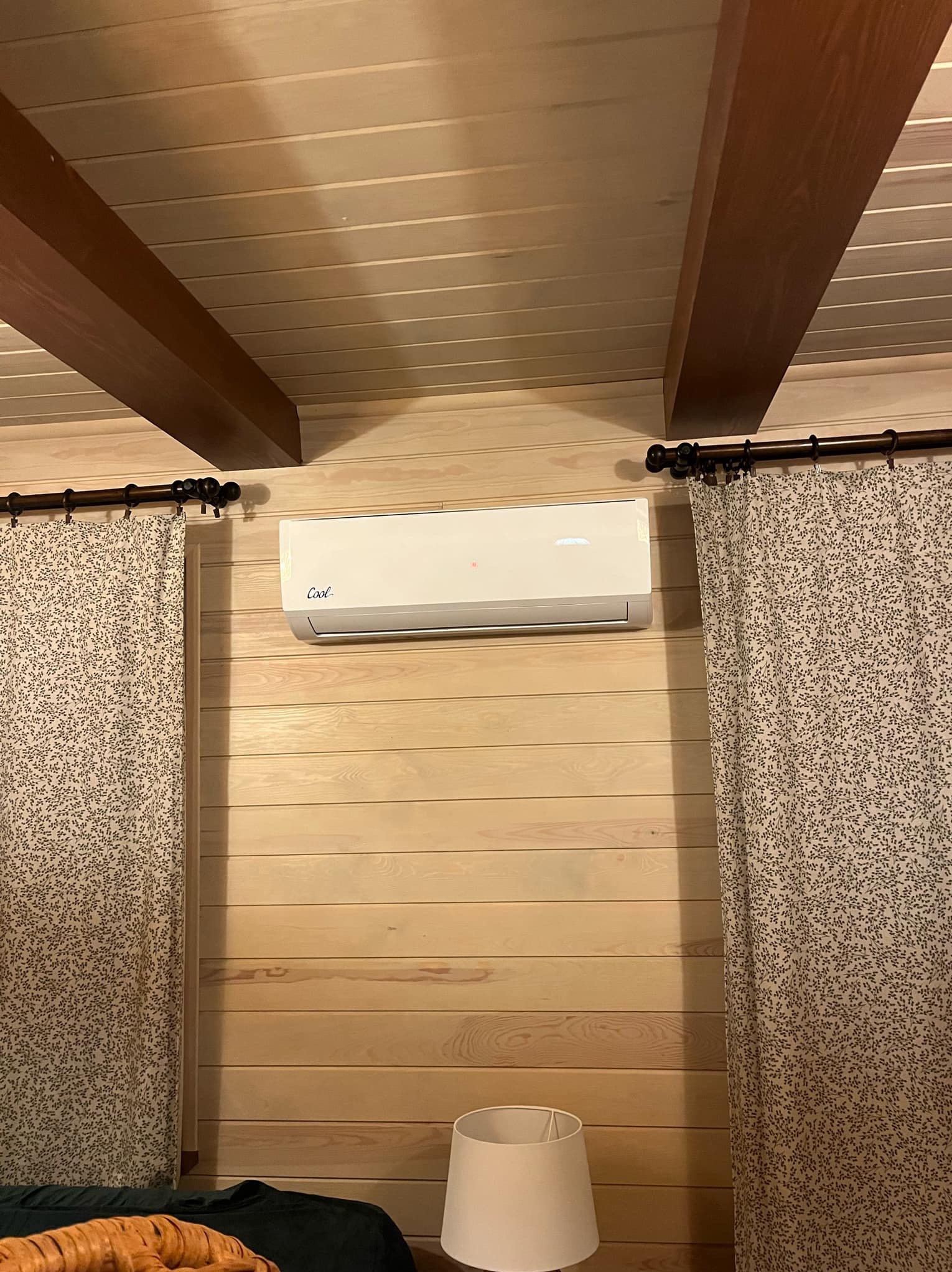 Biały klimatyzator z napisem 'Cool' zamontowany na drewnianej ścianie w pokoju. Widoczne zasłony i fragment lampki nocnej.