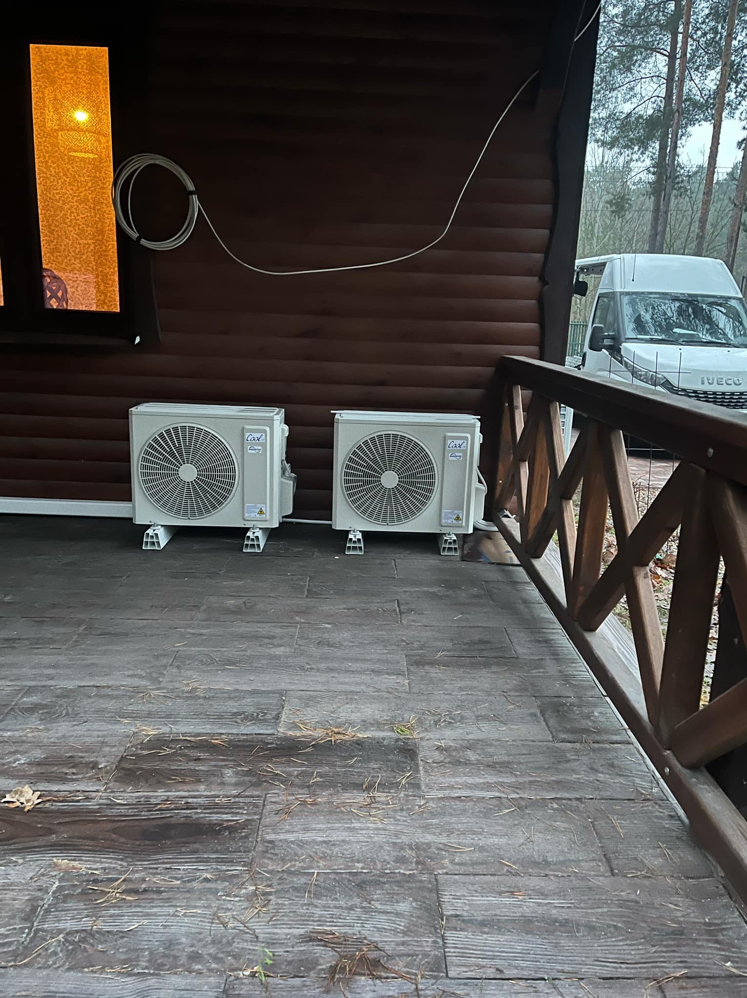 Dwa klimatyzatory na drewnianym tarasie domu z brązową elewacją. Na zewnątrz widoczny biały samochód dostawczy i drzewa.