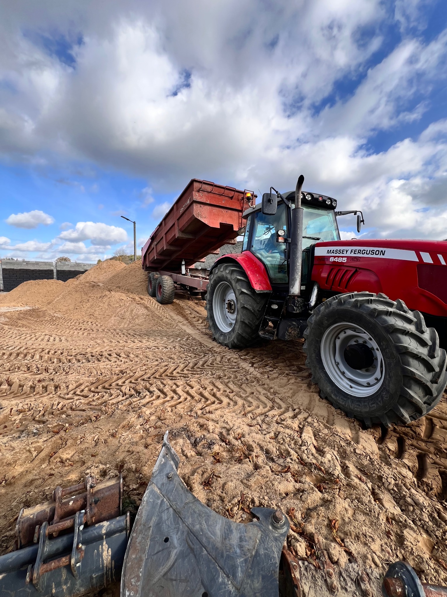 Czerwony traktor Massey Ferguson z przyczepą wywrotką na placu budowy, obok sterta piasku i łyżki koparki, błękitne niebo z chmurami w tle.