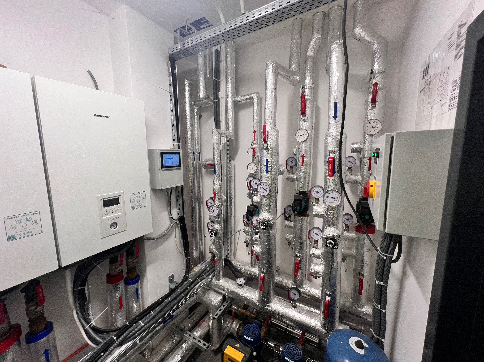 Węzeł cieplny z pompą ciepła Panasonic Aquarea Smart Cloud, licznymi rurami z izolacją, manometrami i zaworami w pomieszczeniu technicznym.
