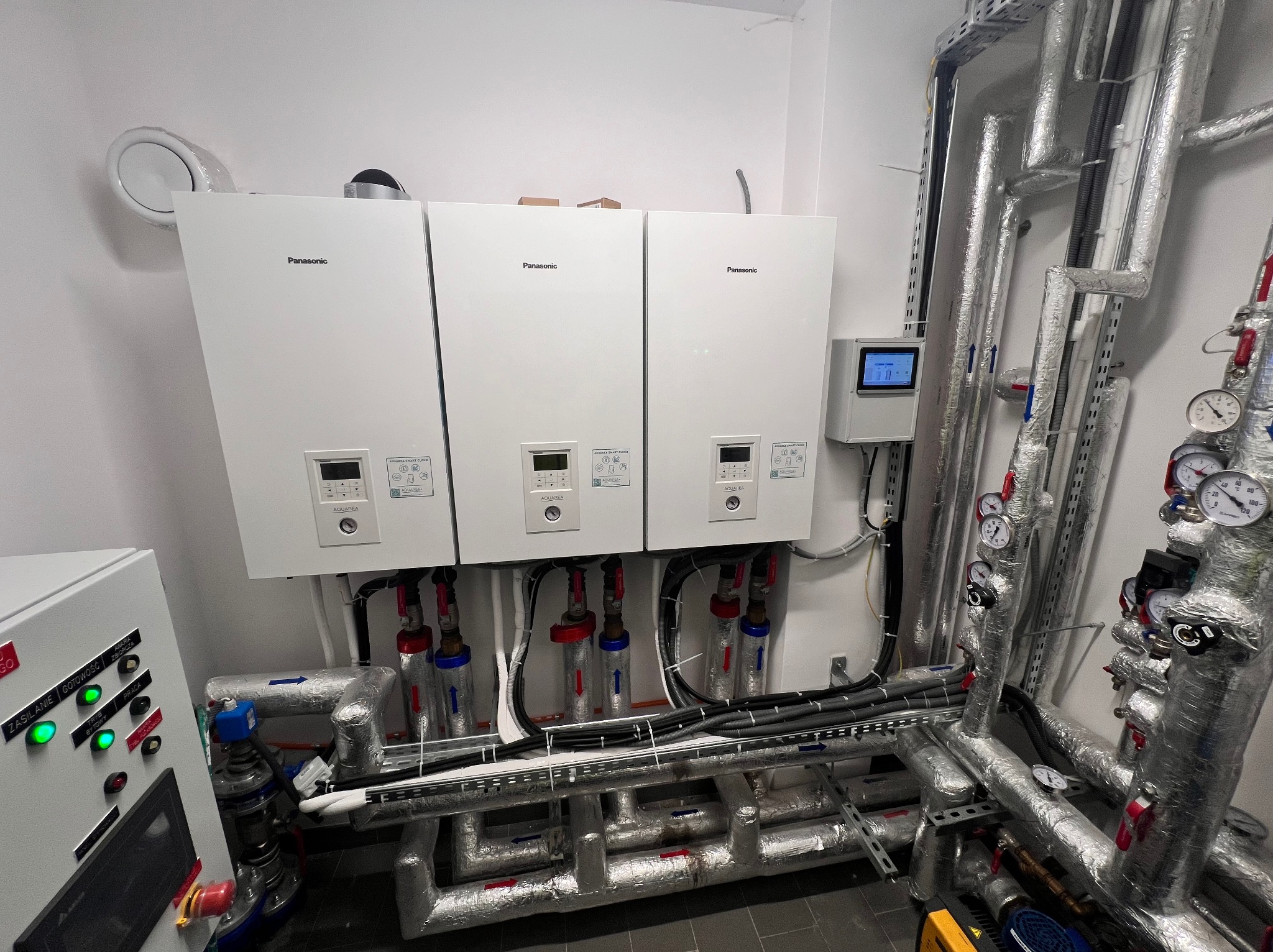 Trzy białe pompy ciepła Panasonic zamontowane na ścianie z rurami, z widocznym panelem sterowania i elementami instalacji. Rury zaizolowane, z manometrami.