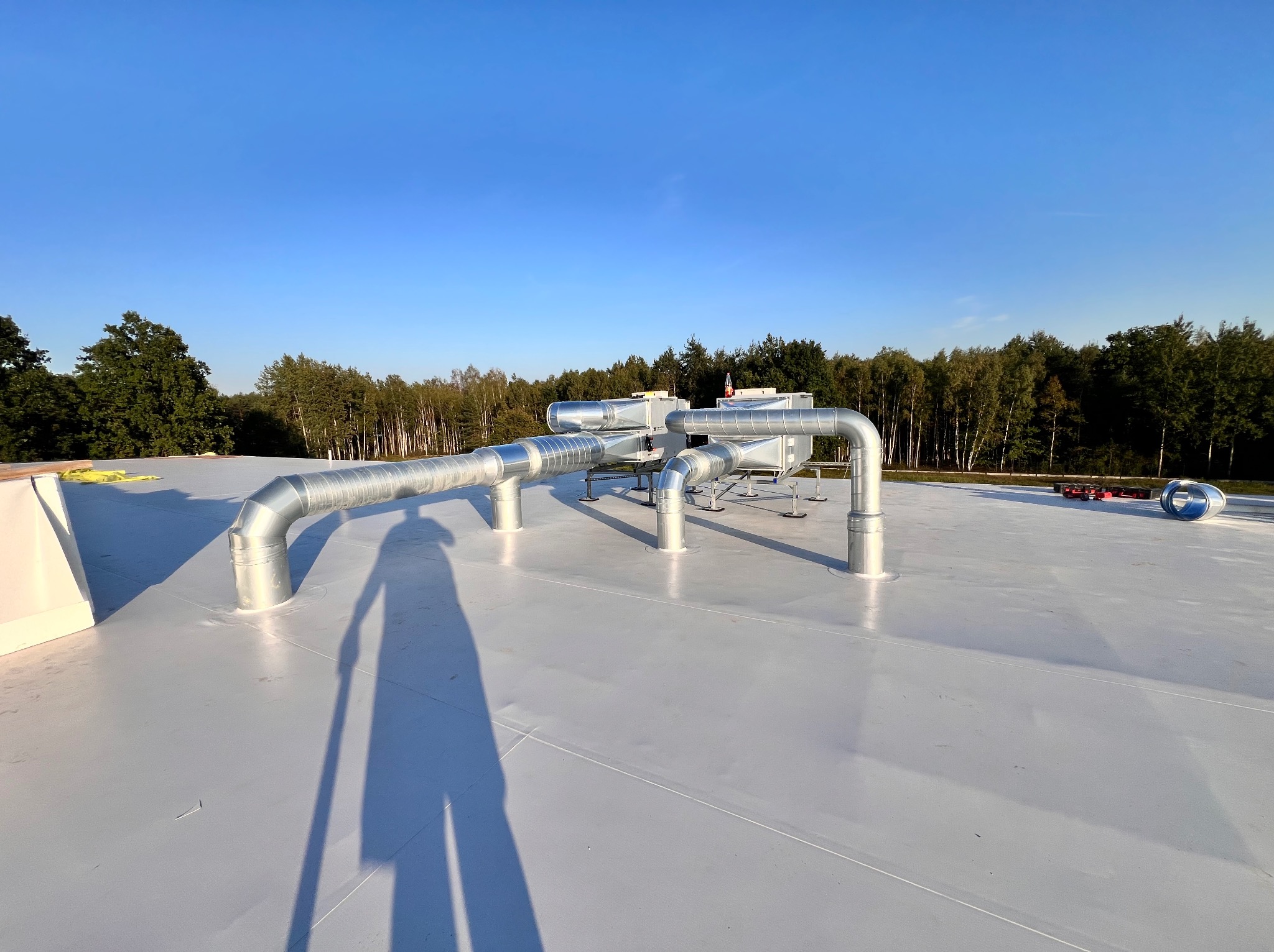 Srebrne rury wentylacyjne na jasnym dachu budynku z widokiem na drzewa i błękitne niebo. Cień osoby widoczny na pierwszym planie.