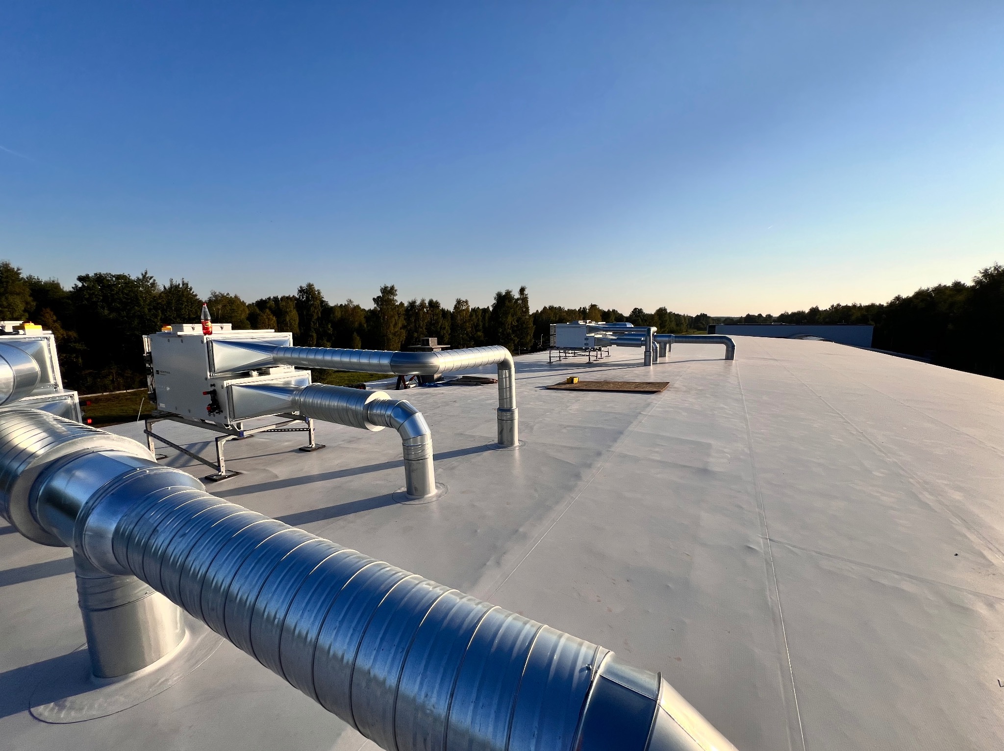 System wentylacyjny z metalowymi rurami na płaskim dachu budynku, widoczne urządzenia i drzewa w tle pod błękitnym niebem.