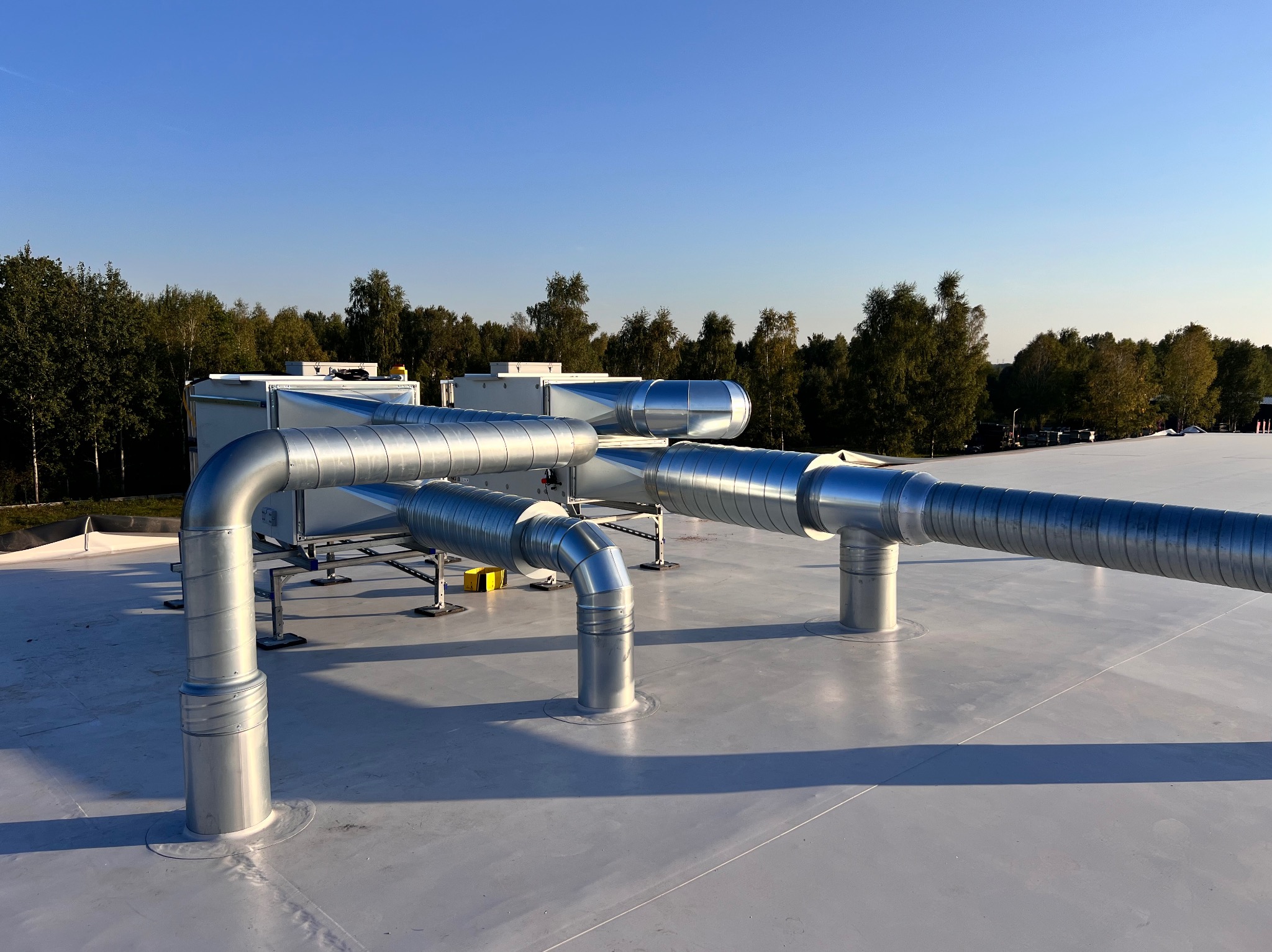 Instalacja wentylacyjna z metalowych rur na dachu budynku, widok z góry na tle zieleni i błękitnego nieba. Rury są srebrne i mają karbowaną strukturę.