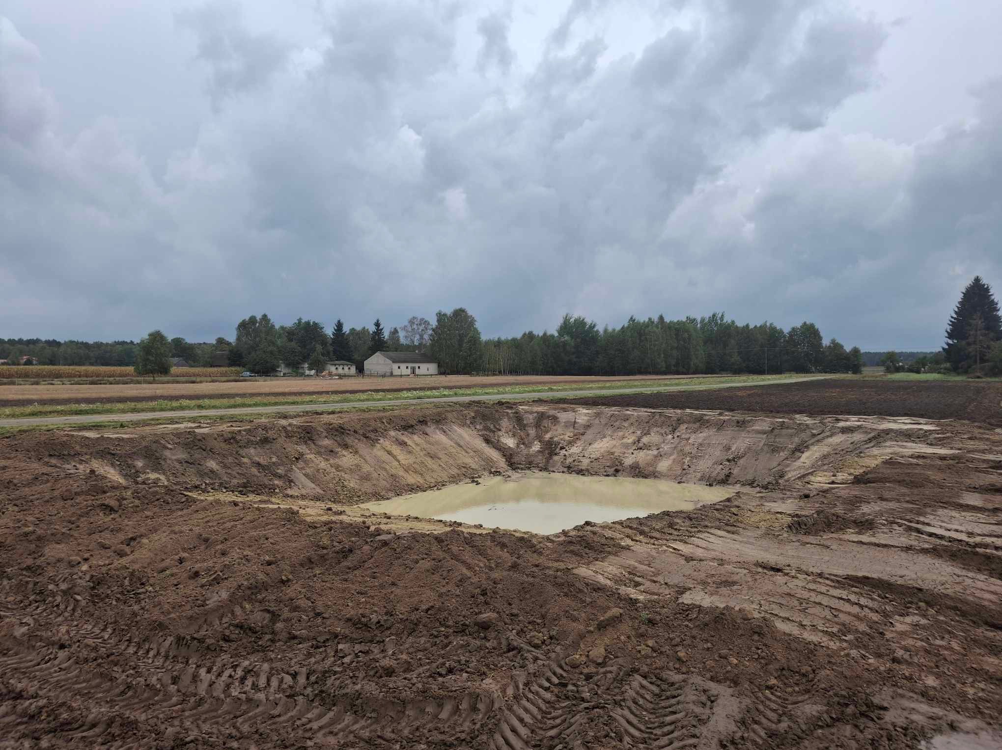 Szeroki wykop w ziemi wypełniony wodą, otoczony polem i lasem pod pochmurnym niebem. Ślady opon na ziemi.