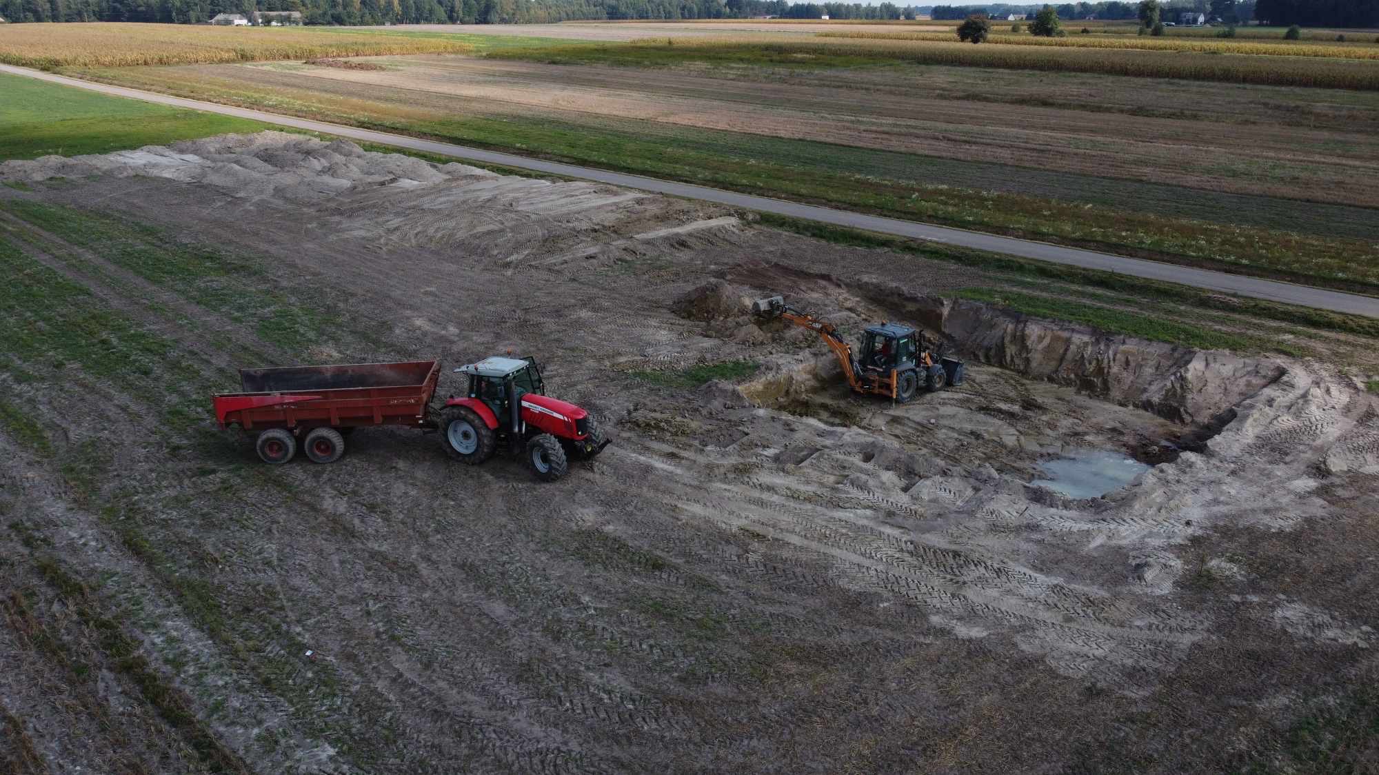 Z lotu ptaka: czerwony traktor z przyczepą i koparko-ładowarka w trakcie prac ziemnych na polu. Wykop pod fundamenty z widocznym zbiornikiem wody.