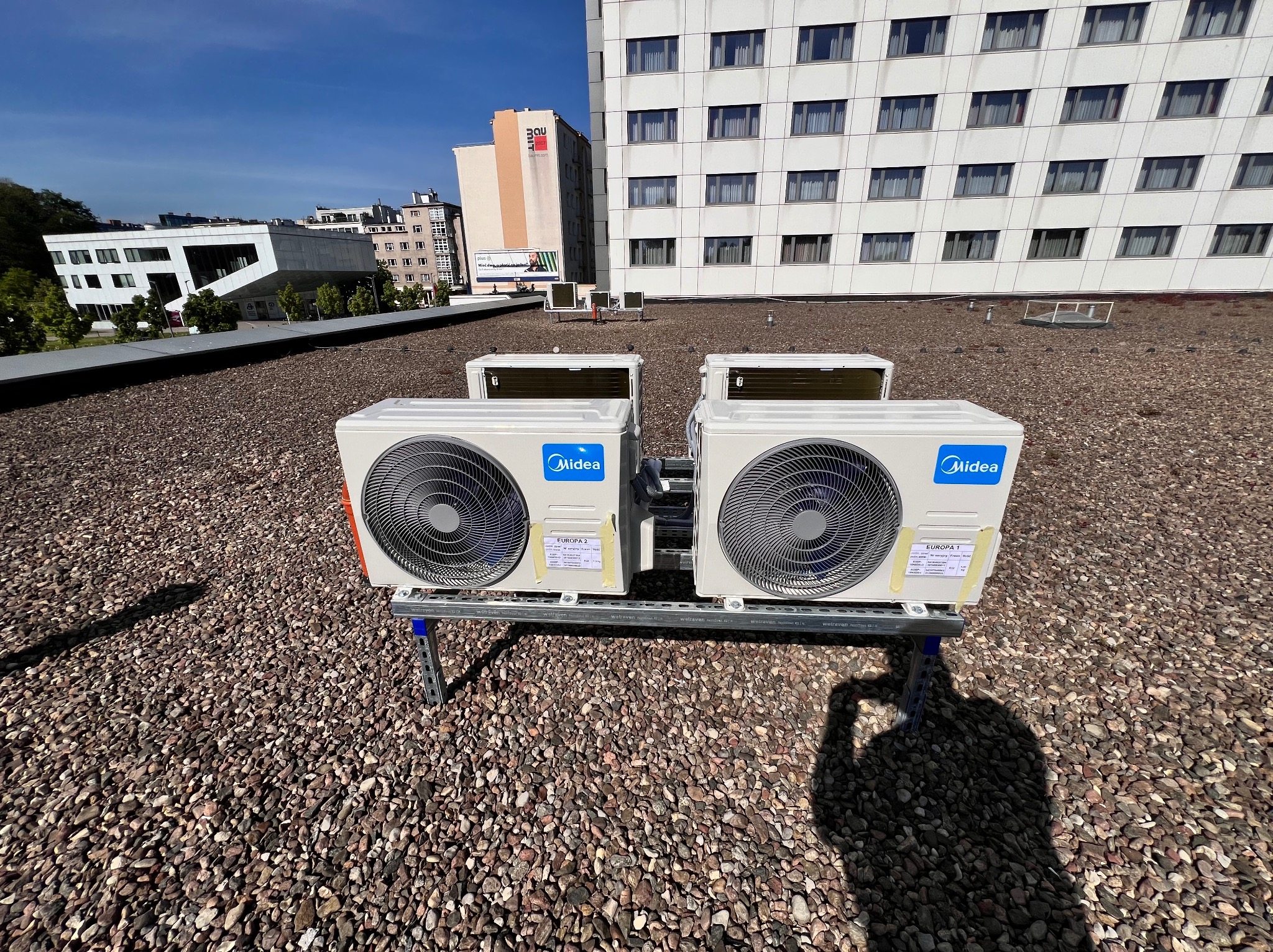 Dwa klimatyzatory Midea, zamontowane na metalowej konstrukcji, na dachu pokrytym drobnym żwirem. W tle budynki w słoneczny dzień. Widoczny cień osoby robiącej zdjęcie.