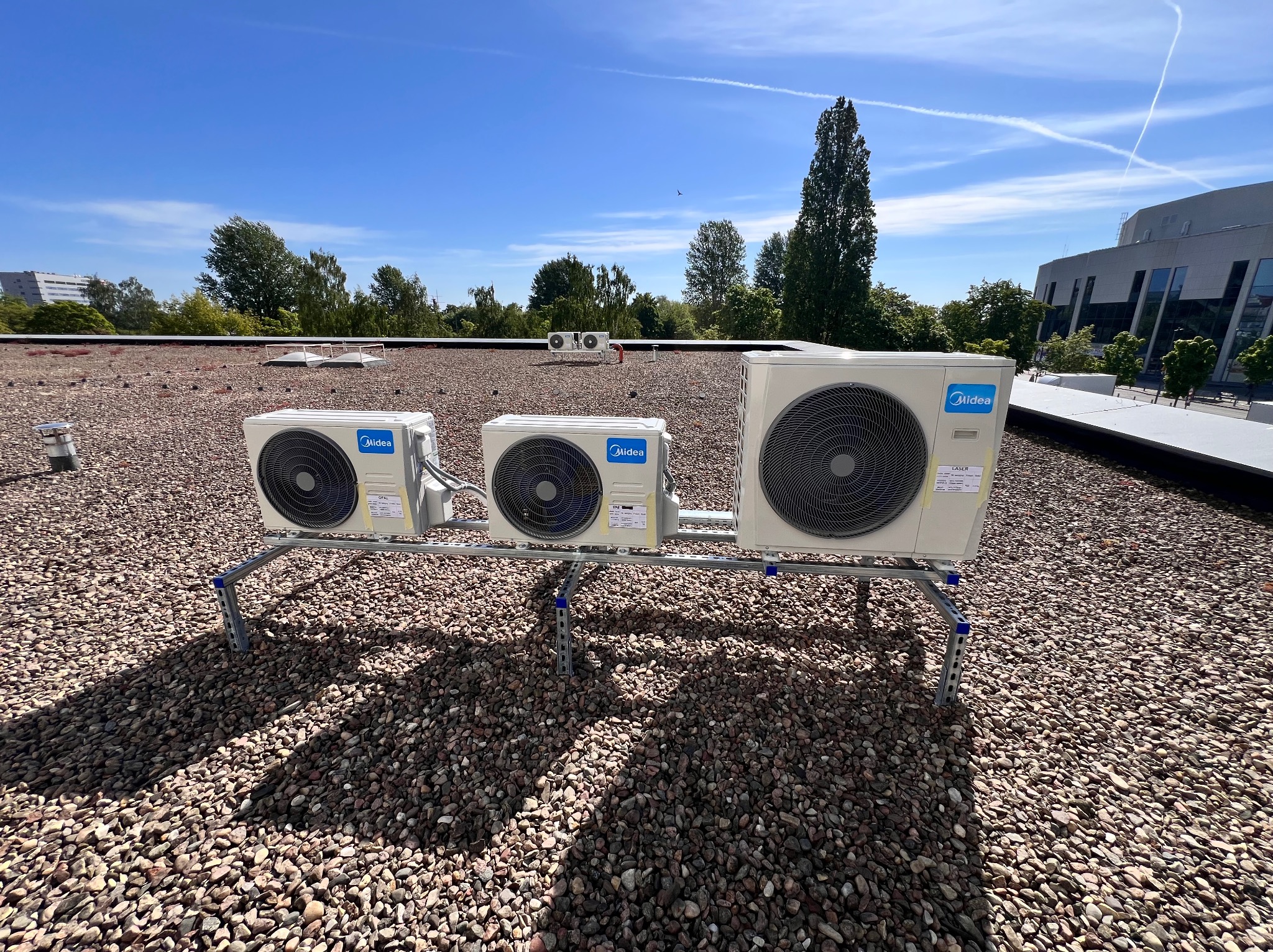 Zestaw klimatyzatorów Midea na dachu pokrytym żwirem, zamontowanych na metalowej konstrukcji. W tle budynek i błękitne niebo z białymi smugami kondensacyjnymi.