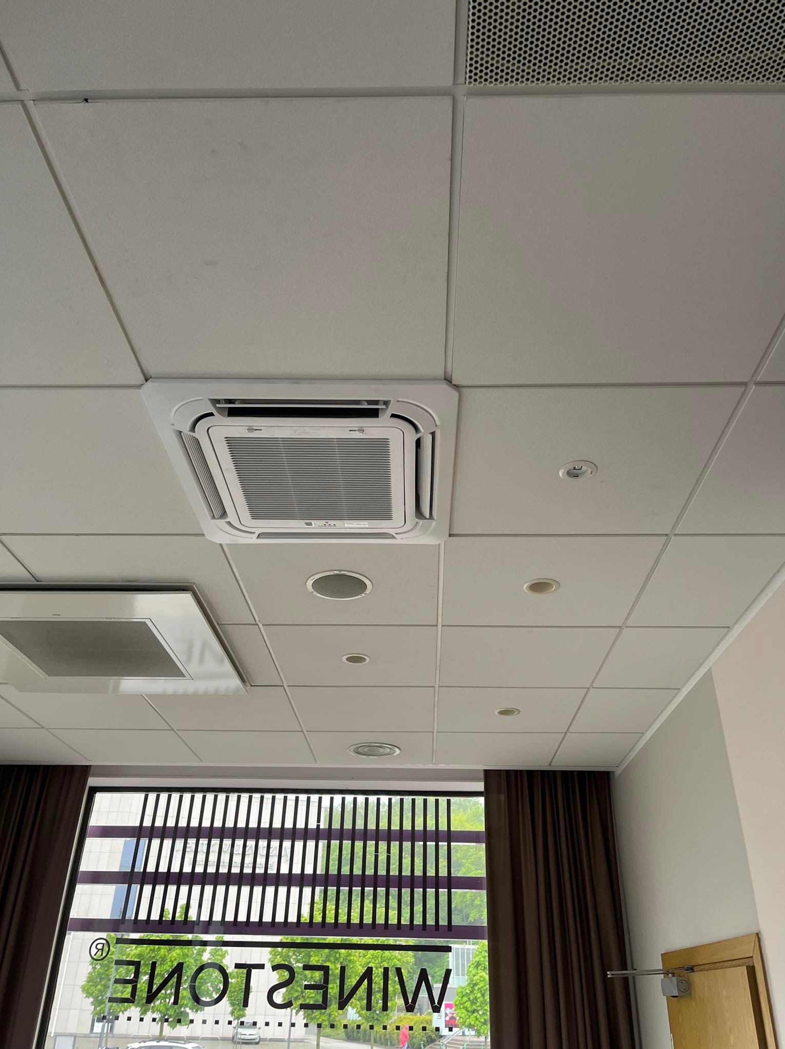 Kasetonowa klimatyzacja sufitowa z widoczną kratką wentylacyjną i oświetleniem punktowym. Okno z żaluzjami i widokiem na zieleń.