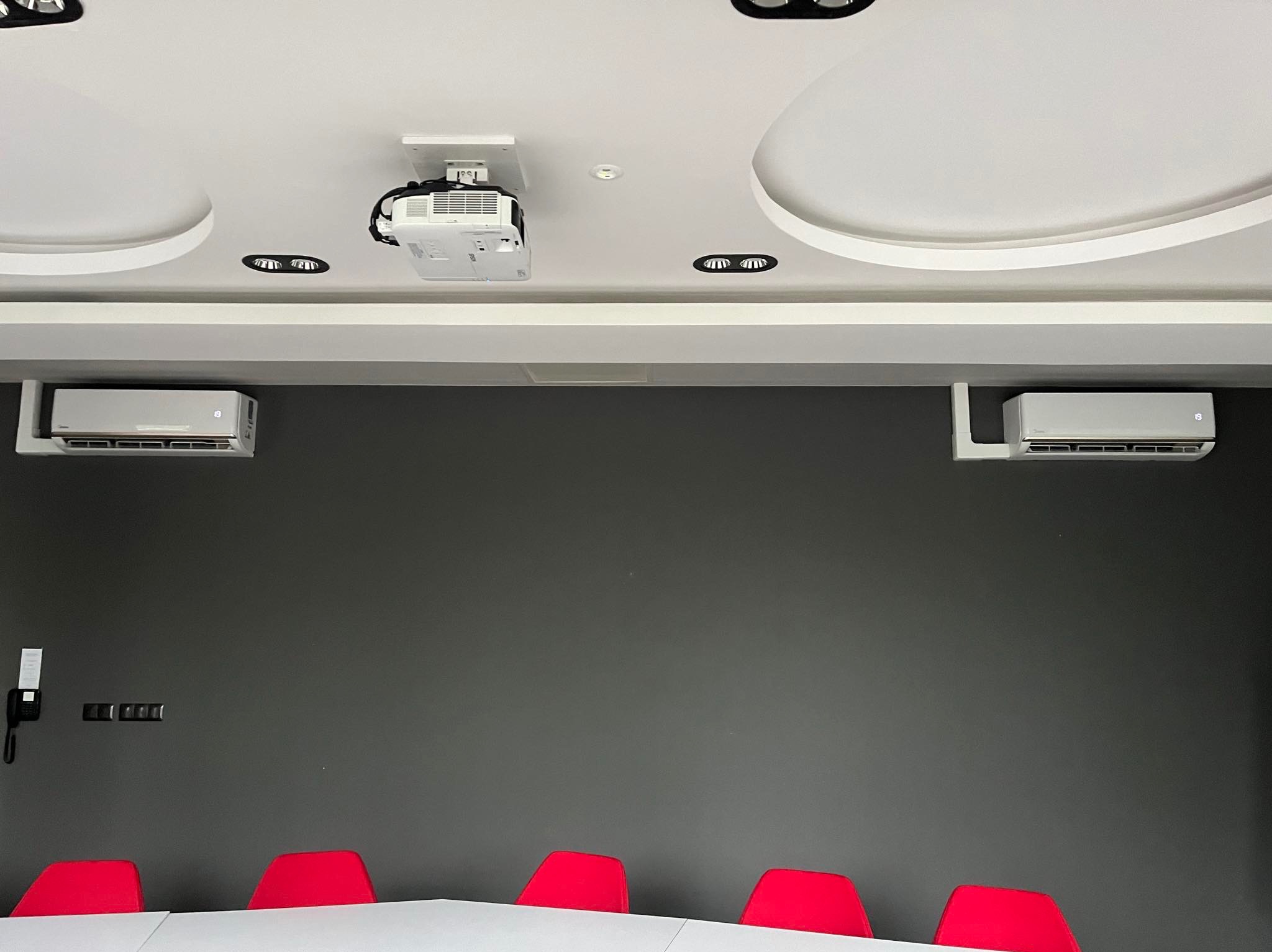 Klimatyzacja w biurze: Dwa białe klimatyzatory ścienne Midea, projektor sufitowy i czerwone krzesła konferencyjne na tle szarej ściany.