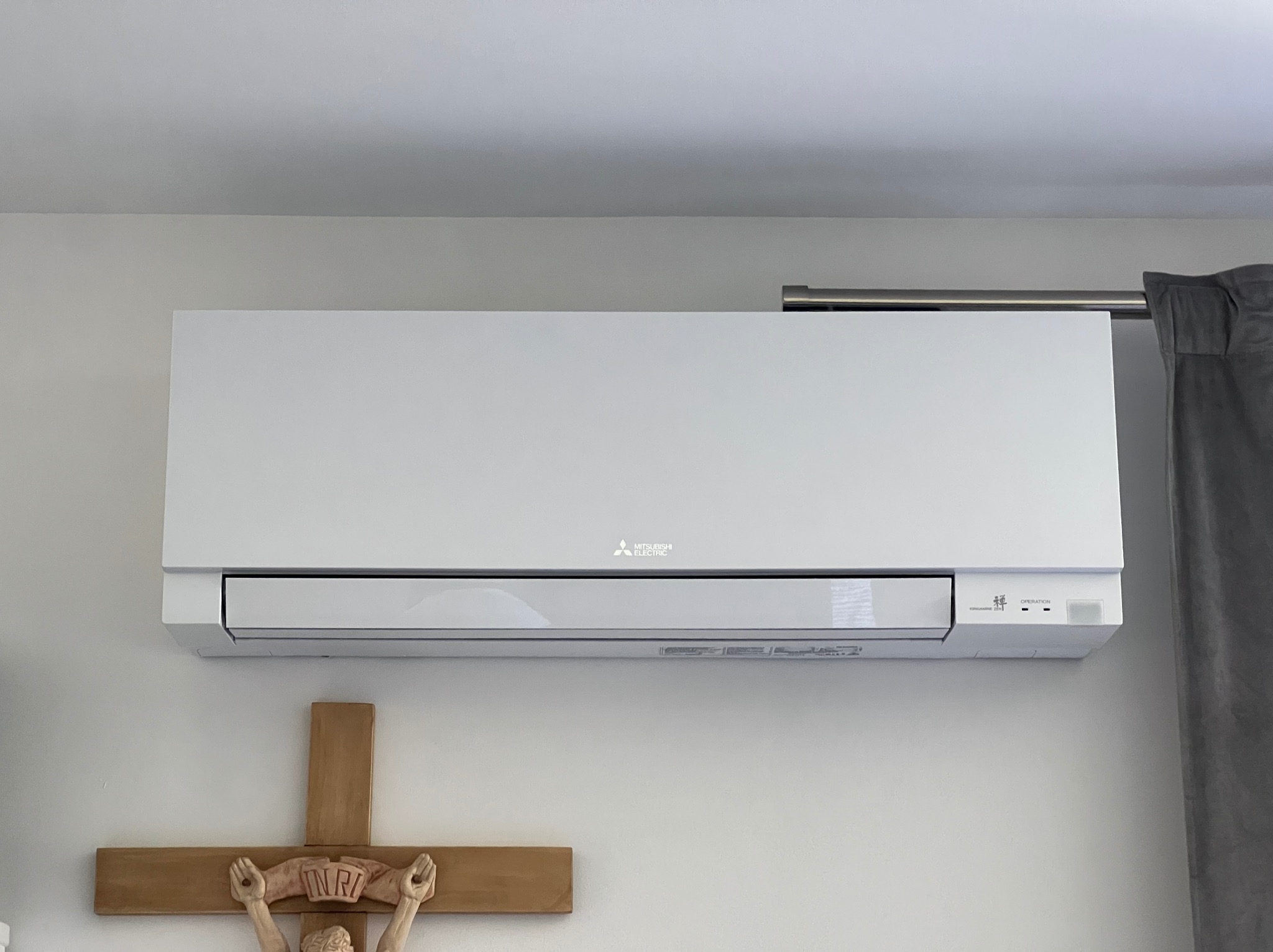 Nowoczesny, biały klimatyzator Mitsubishi Electric zamontowany na jasnej ścianie, widoczny fragment karnisza z zasłoną i krzyż z Jezusem na ścianie.