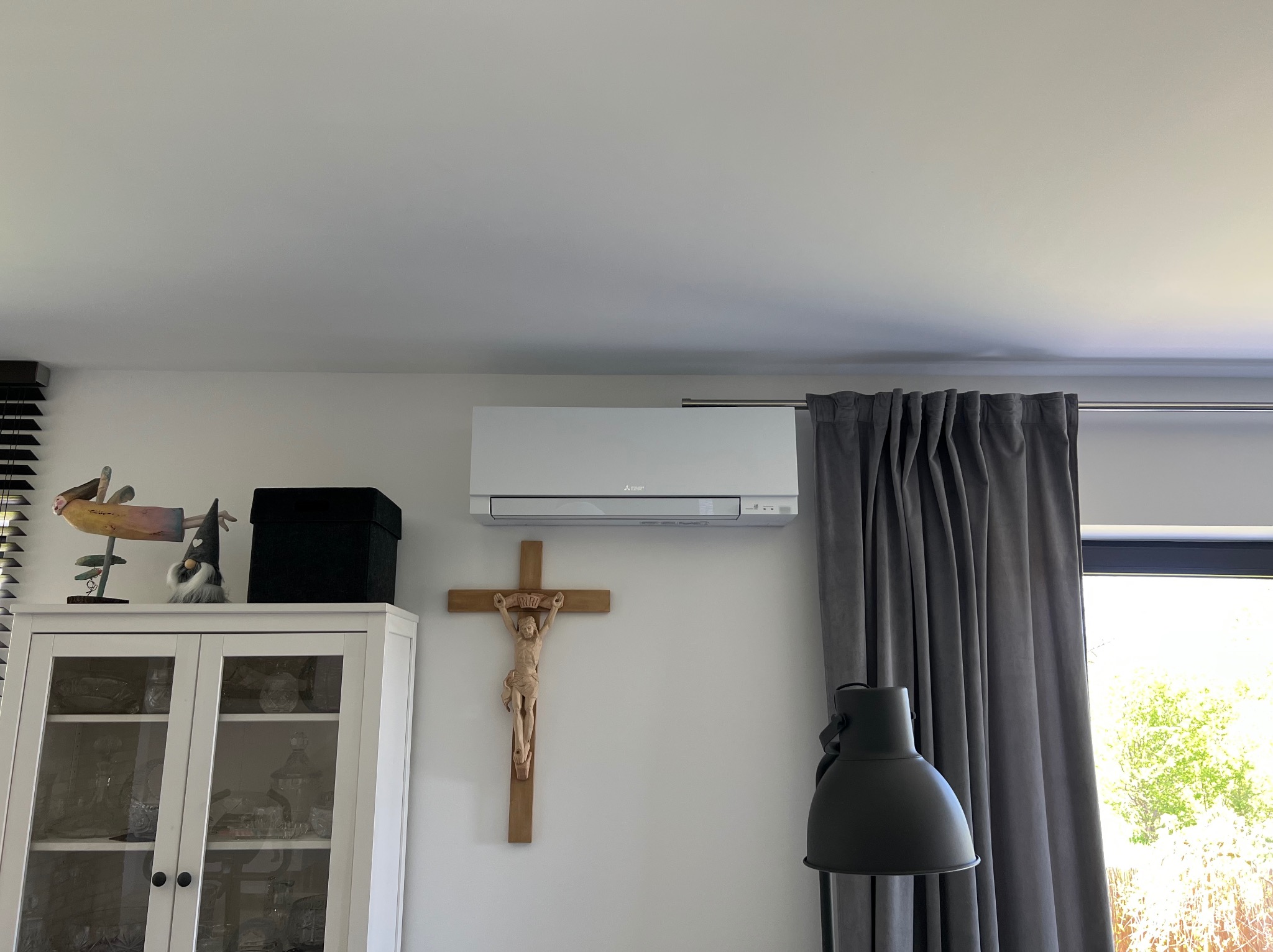 Nowoczesny klimatyzator Mitsubishi zamontowany na ścianie nad drewnianym krzyżem w jasnym wnętrzu, obok okna z szarymi zasłonami i lampą.