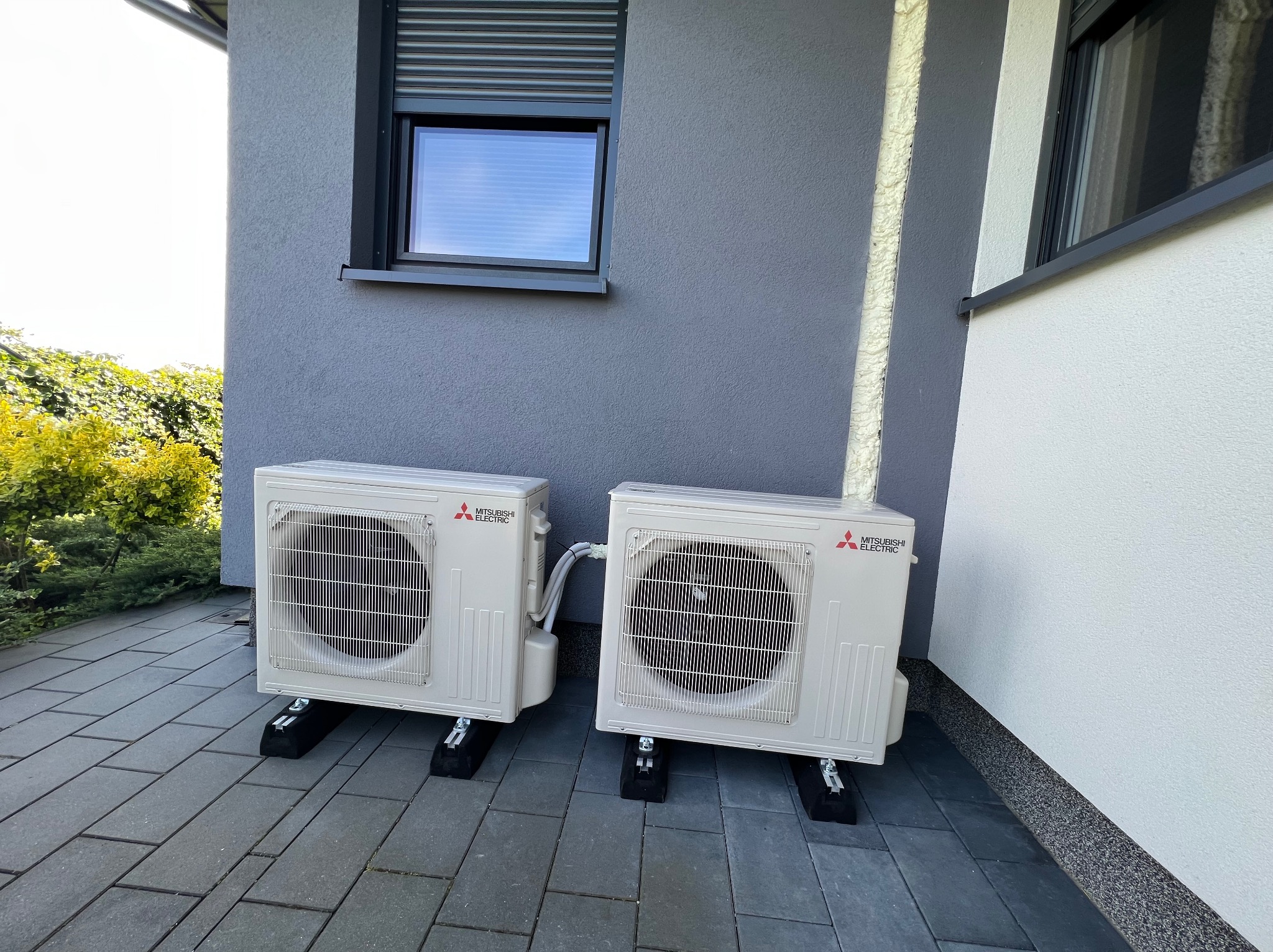 Dwa białe klimatyzatory Mitsubishi Electric na czarnych podstawach, umieszczone na szarej kostce brukowej przy ścianie budynku z oknem z roletą zewnętrzną.