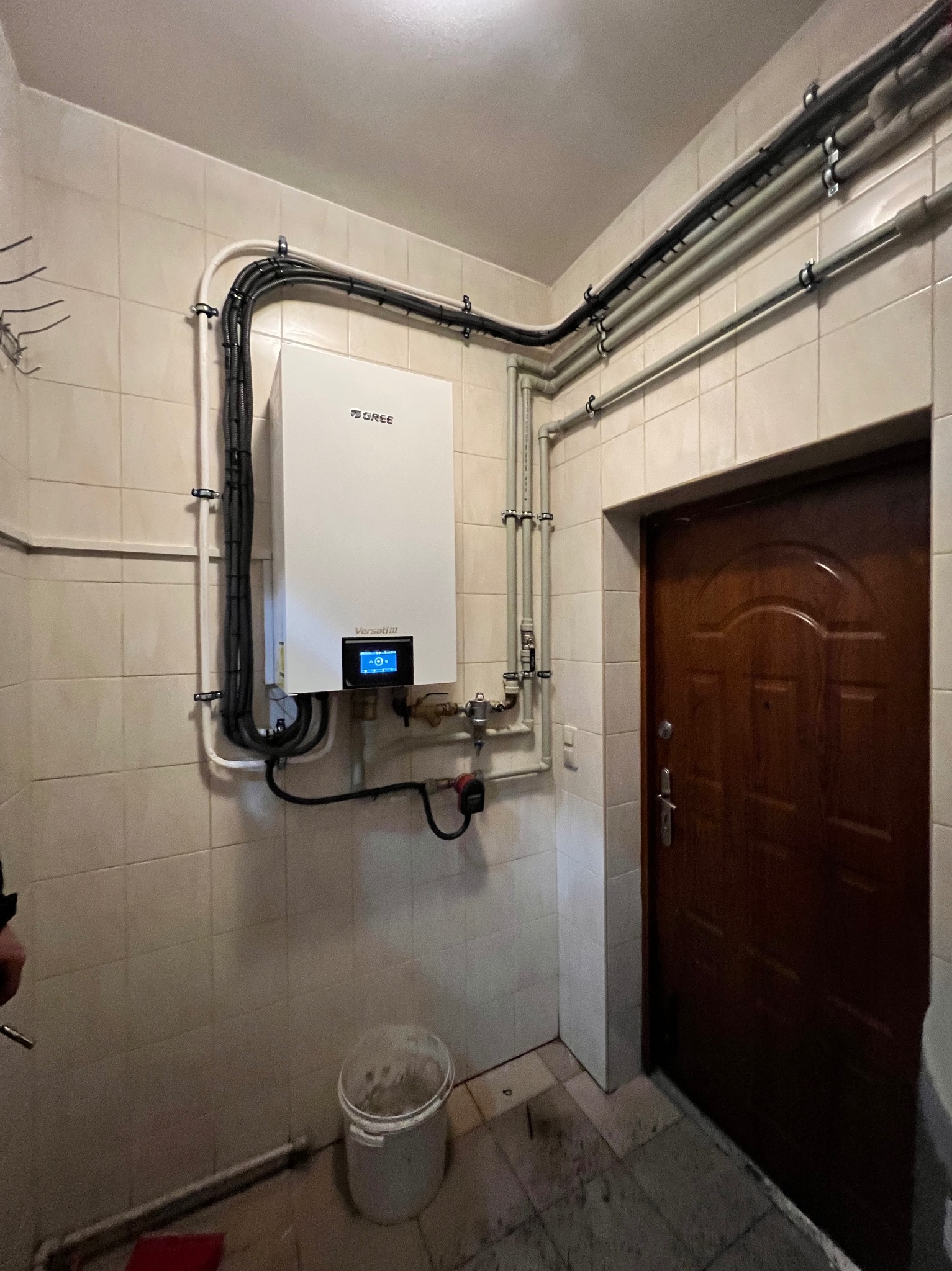 Zamontowany biały piec grzewczy marki Gree na kafelkowej ścianie, z widocznymi rurami i przewodami, w pomieszczeniu z brązowymi drzwiami i wiadrem na podłodze.