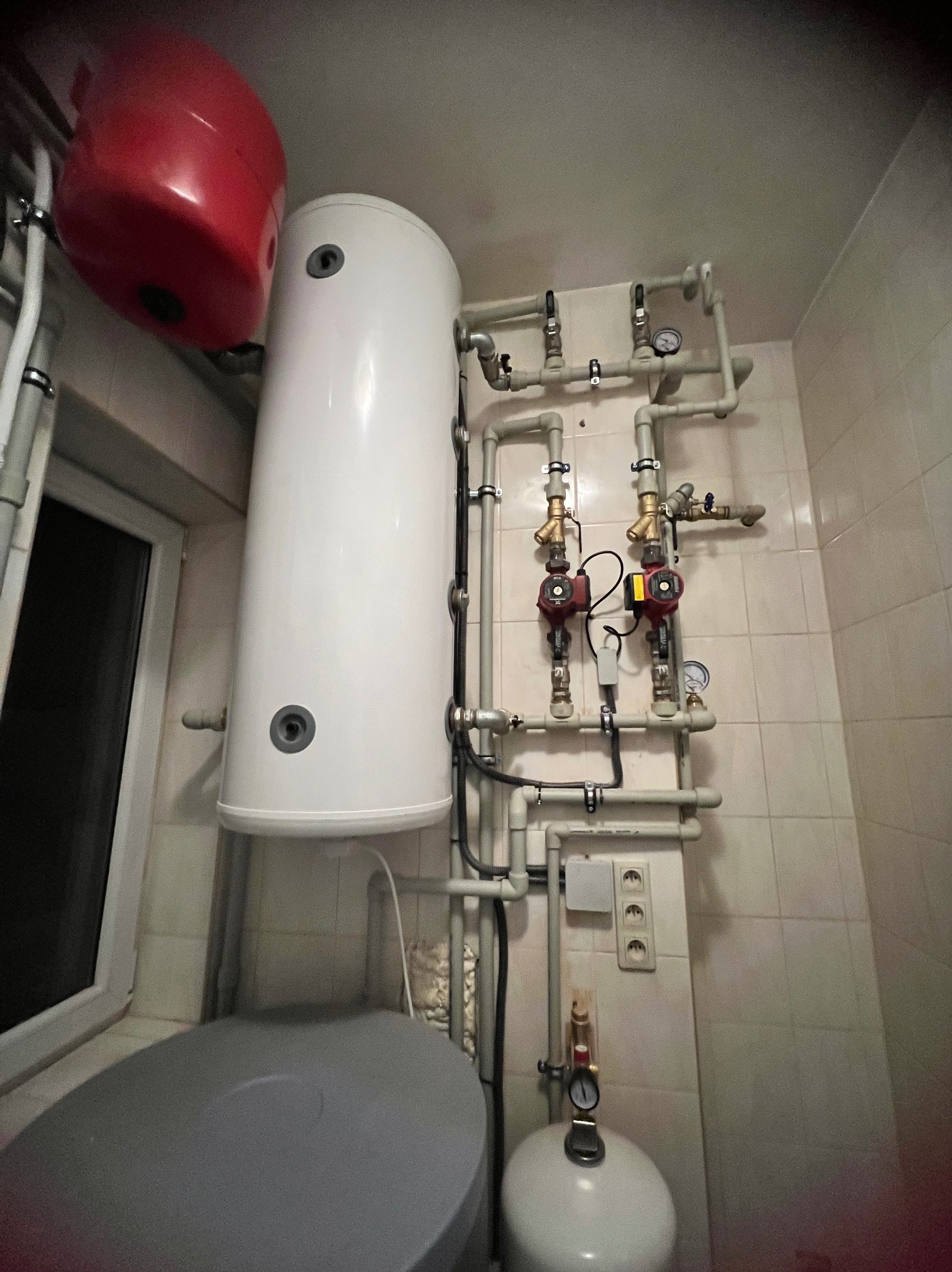 Biały, pionowy zbiornik buforowy z czerwonym naczyniem wzbiorczym i pompami obiegowymi w kotłowni z białymi płytkami. Instalacja grzewcza w pomieszczeniu.