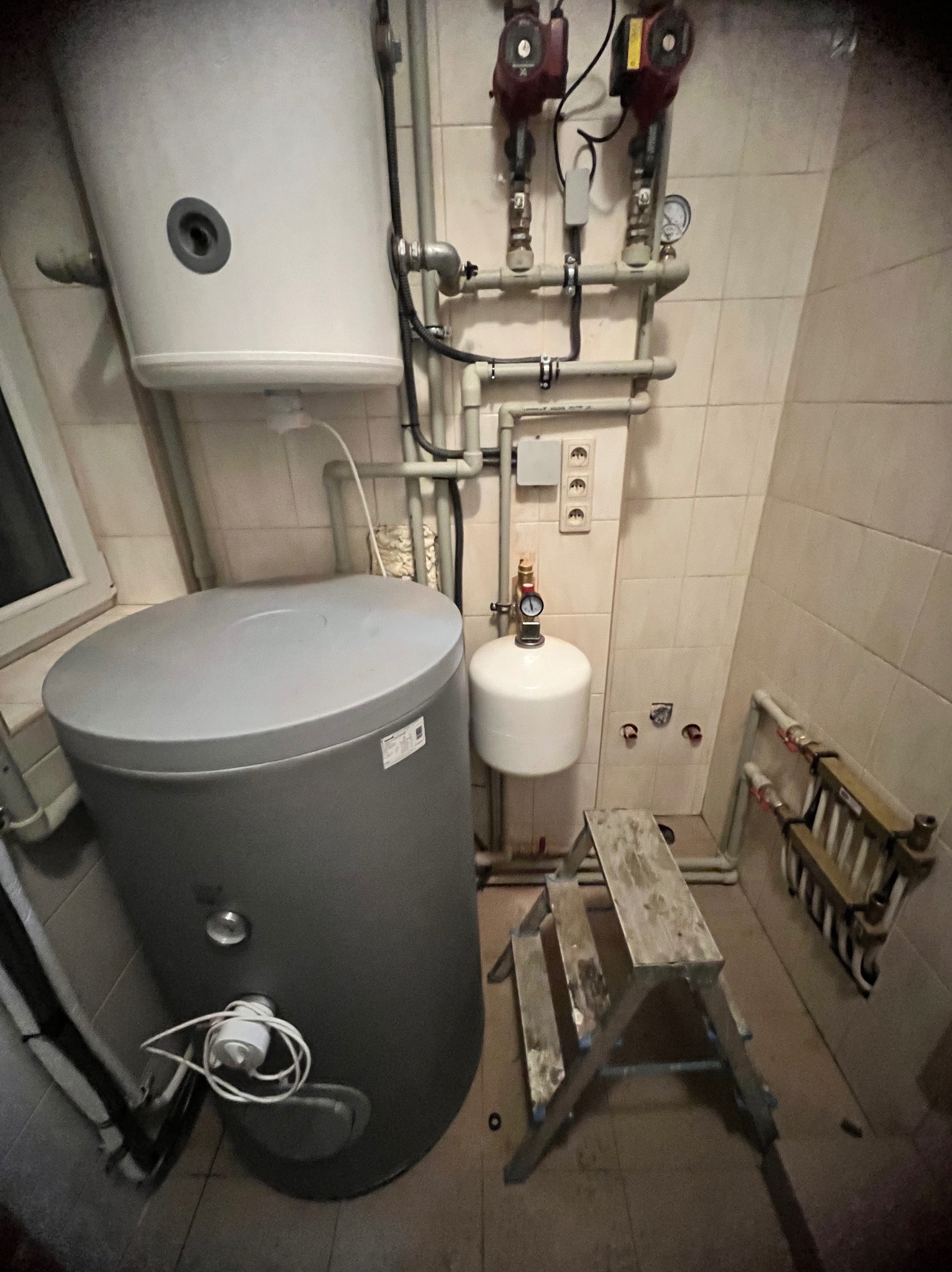 Szary zbiornik buforowy z białym zasobnikiem wody i instalacją hydrauliczną na tle beżowych płytek. Obok drabina.