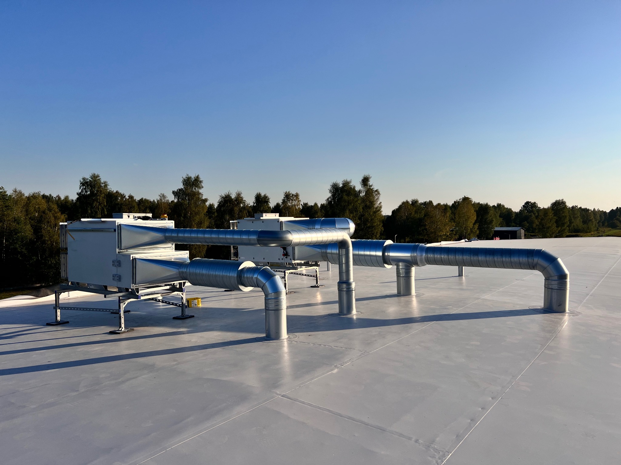 Kompletny system wentylacyjny z metalowymi rurami na jasnym dachu budynku, z drzewami w tle i błękitnym niebem. Widok z dołu na instalację.