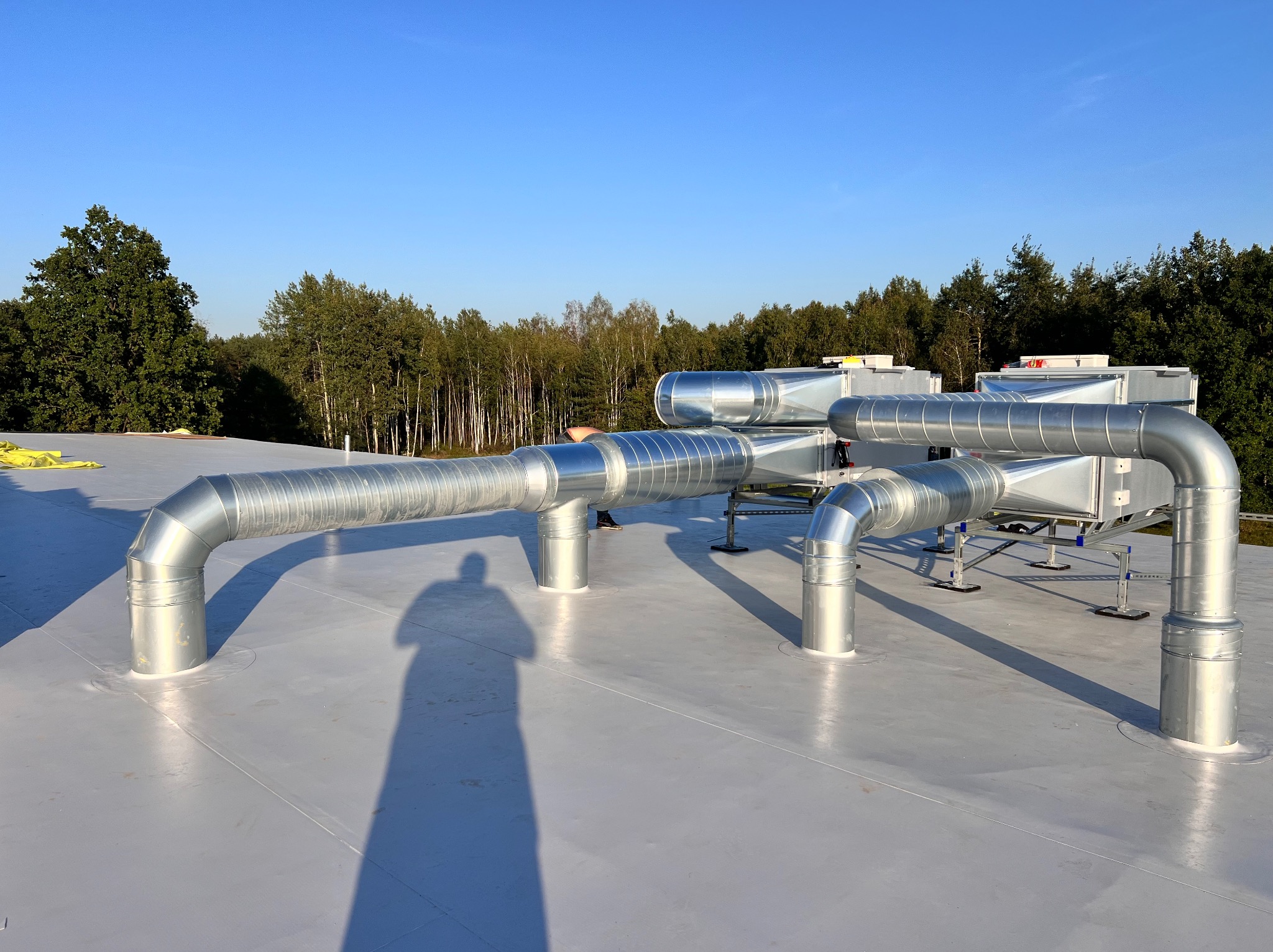 Kompleksowa instalacja wentylacyjna na dachu budynku w słoneczny dzień. Widoczne metalowe rury i urządzenia wentylacyjne. Cień osoby w kadrze.