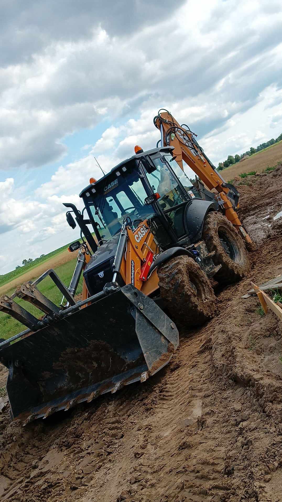 Pomarańczowa koparko-ładowarka CASE w błotnistym terenie, widok z dołu, ślady opon w ziemi, pola w tle pod pochmurnym niebem.