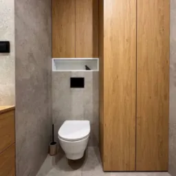 Nowoczesna łazienka z szarą płytką, zabudową z drewna i podwieszaną toaletą. Minimalistyczny design z akcentami czerni i dekorem w postaci figurki ptaka.