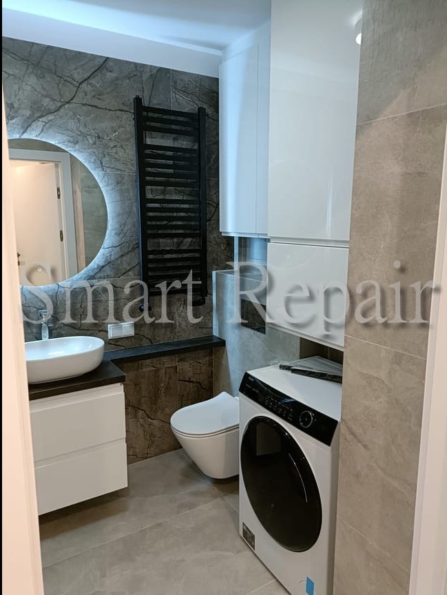 Nowoczesna łazienka z umywalką, toaletą, pralką i grzejnikiem na tle marmurowych płytek, oświetlona okrągłym lustrem z podświetleniem LED.