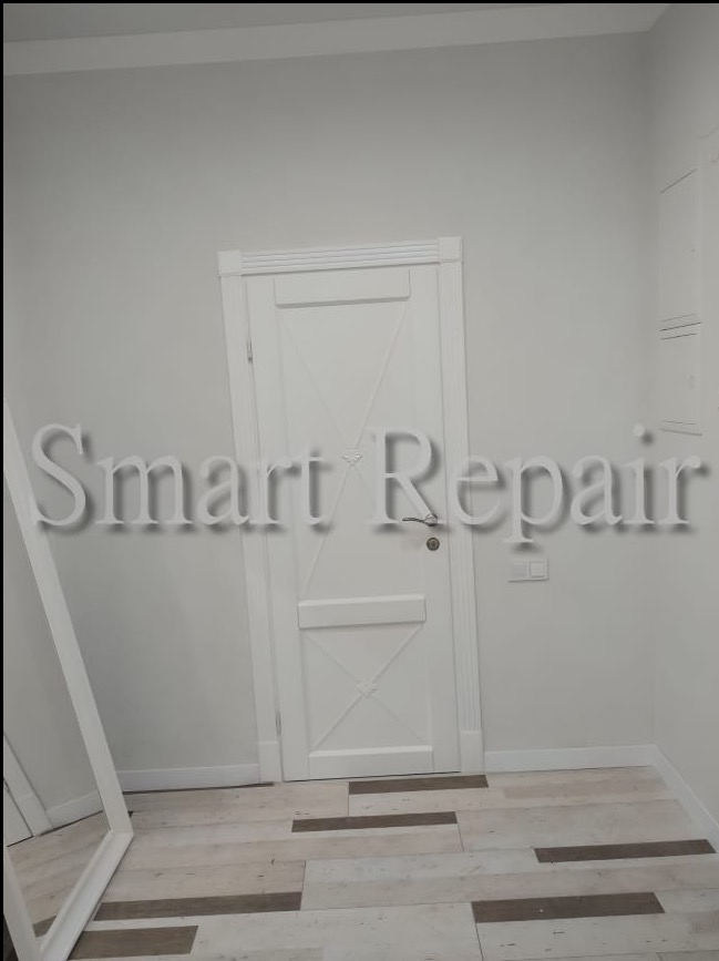 Białe, odnowione wnętrze: drzwi z dekoracyjnym wzorem, jasne ściany i podłoga imitująca drewno. Widoczne gniazdko elektryczne i fragment lustra po lewej stronie.